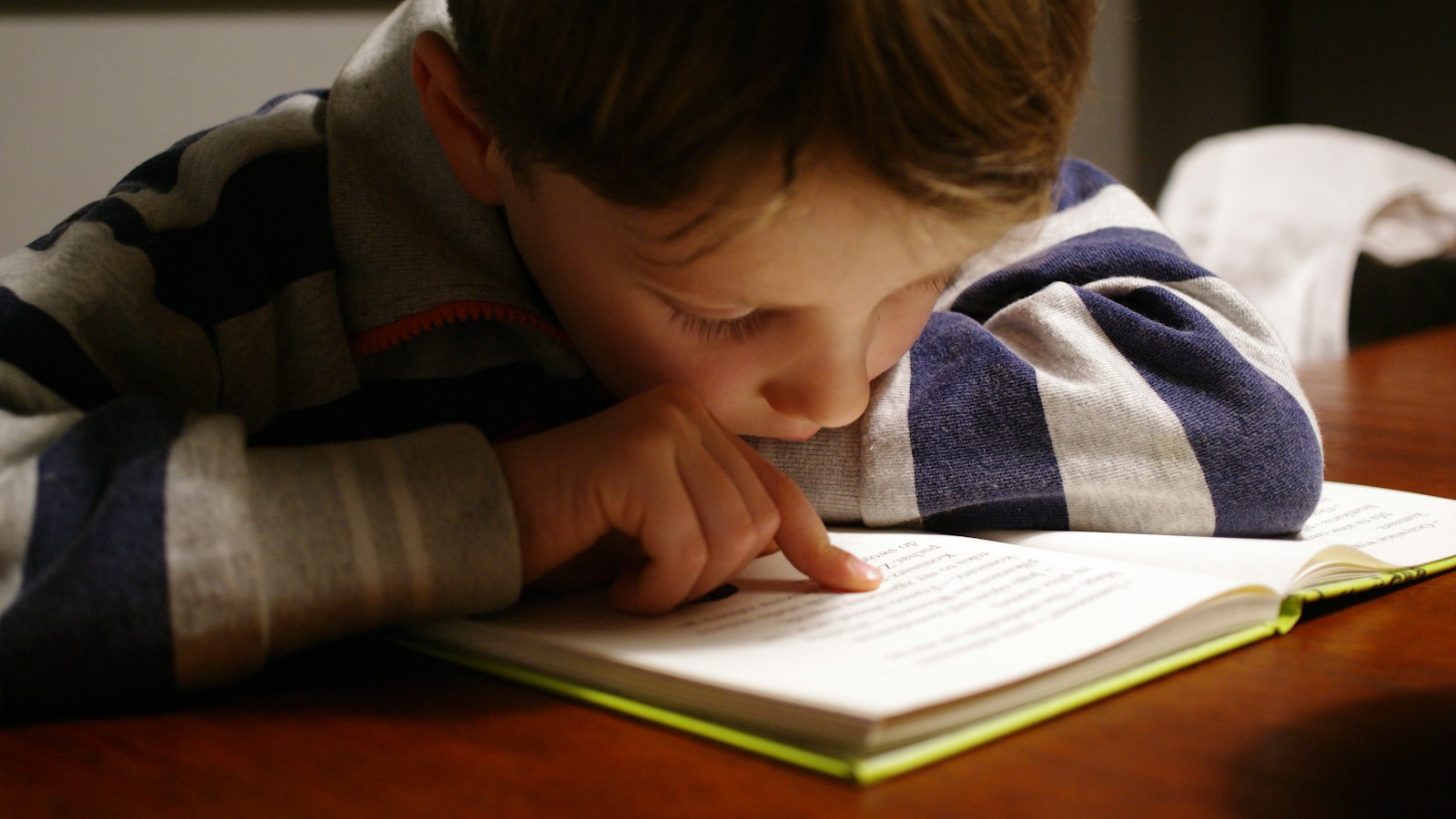 A child in a striped shirt reading a book at a wooden table