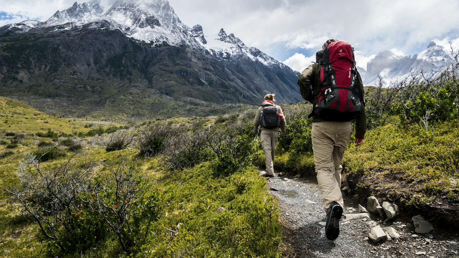 Two hikers with backpacks walking along a trail toward snow-capped mountains