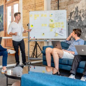 three men sitting while using laptops and watching man beside whiteboard