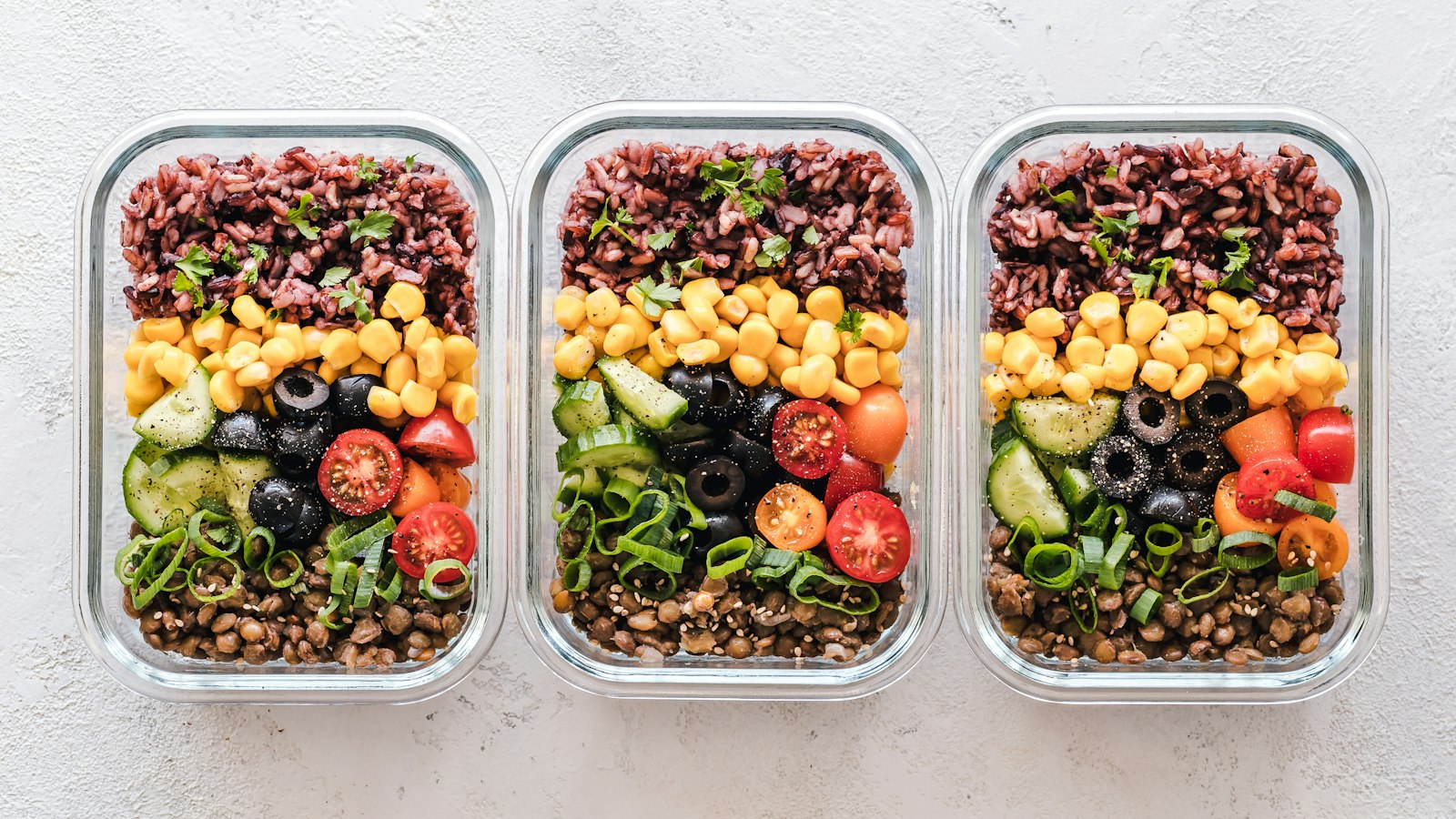 Three glass containers filled with rice, corn, olives, tomatoes, cucumbers, and lentils