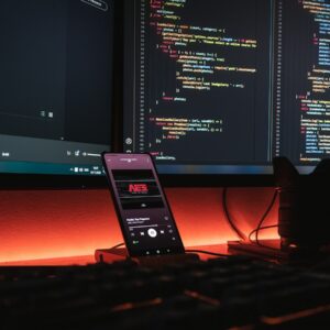 A smartphone displaying music on a desk with computer monitors showing code