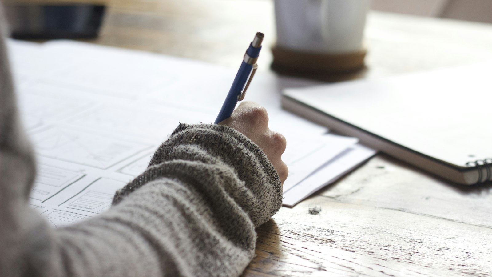person writing on brown wooden table near white ceramic mug